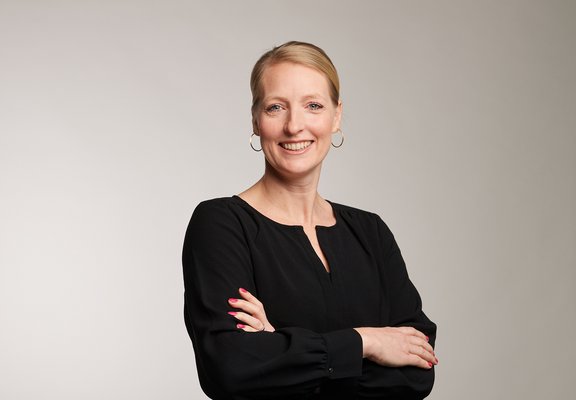 Auf dem Bild sieht man Johanna Ebbing. Johanna leitet den Nachhaltigkeitsbereich, das Employee Tech Team sowie das Facility Team. Sie hat blonde Haare, trägt ein schwarzes Kleid und lächelt in die Kamera.  