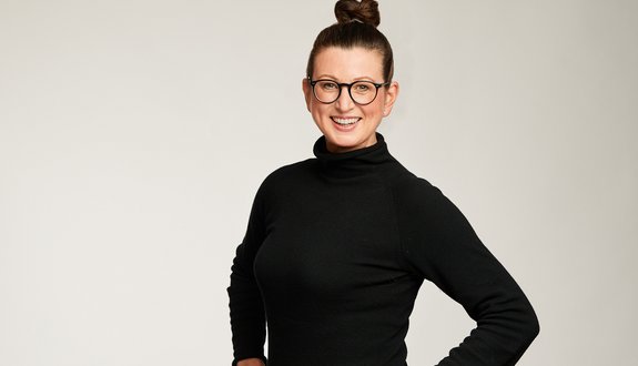 Auf dem Bild ist Dr. Claudia Viehweger, die Chief Sustainability Officer von Scout24 zu sehen. Sie hat die Haare in einen Dutt gebunden, trägt eine Brille und ein schwarzes Kleid. Sie lächelt in die Kamera. 