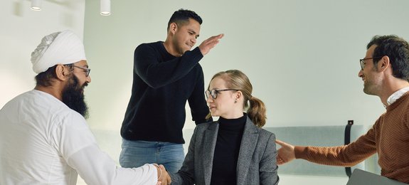 Vier Mitarbeitende sind zusammen in einem Büro. Links ein Mann mit Turban, daneben ein Mann in dunklem Pullover, eine Frau mit blonden Haaren in grauem Blazer und rechts ein Mann in braunem Pullover. Sie befinden sich in lockerer Gesprächsrunde vor einer hellen Wand.