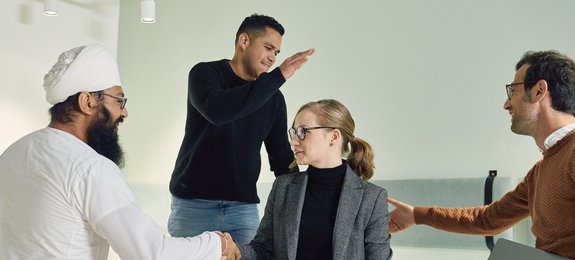Four employees are together in an office. On the left, a man wearing a turban, next to him a man in a dark sweater, a woman with blonde hair in a gray blazer, and on the right a man in a brown sweater. They are engaged in a casual conversation in front of a light wall.