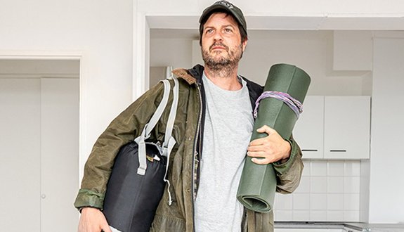 A man with a beard and a baseball cap stands in an empty apartment. He is wearing a green outdoor jacket, holding a rolled-up sleeping mat under his right arm and carrying a large black bag over his left shoulder.
