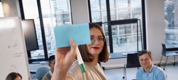In a modern office, an employee with glasses and brown hair holds a blue note and a pen. She is wearing a light-colored top and smiling. Other employees are seated in the background.
