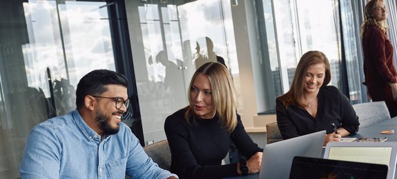 Drei Mitarbeitende sitzen in einem modernen Büro mit großen Fenstern zusammen. Links sitzt ein Mann mit Brille am Laptop, daneben eine Frau mit blonden Haaren, rechts eine Frau mit dunklen Haaren. Im Hintergrund steht eine weitere Person.