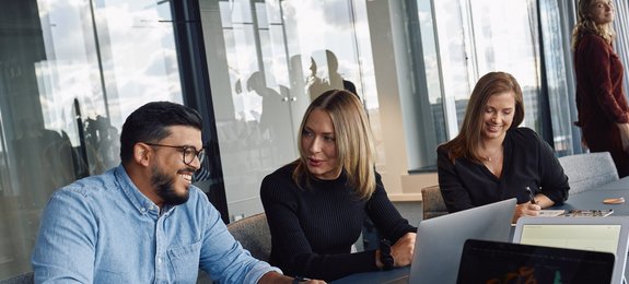 Three employees are sitting together in a modern office with large windows. On the left, a man with glasses is working on a laptop, next to him a woman with blonde hair, and on the right a woman with dark hair. Another person is standing in the background.